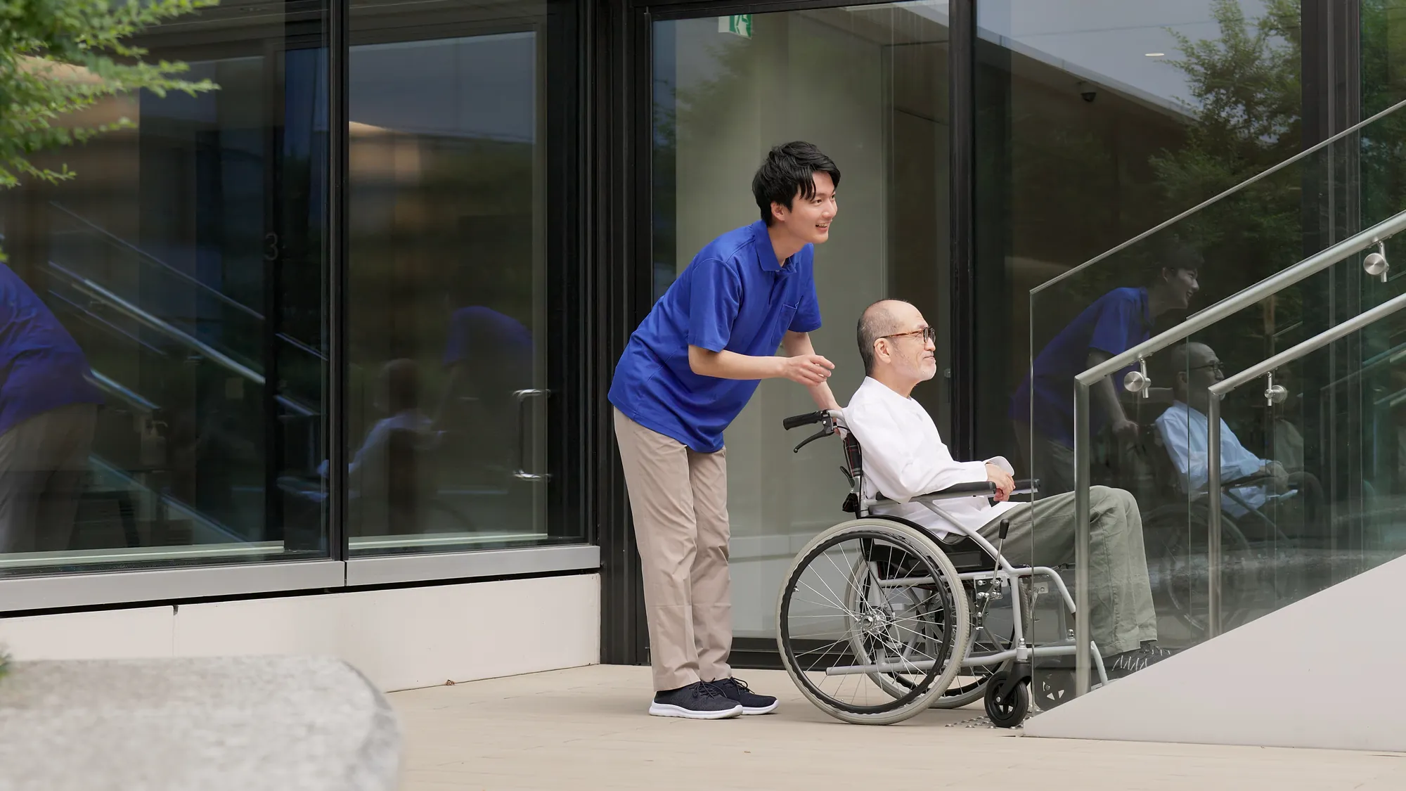 Nurse leaning in to talk with a senior man in a wheelchair at a building entrance