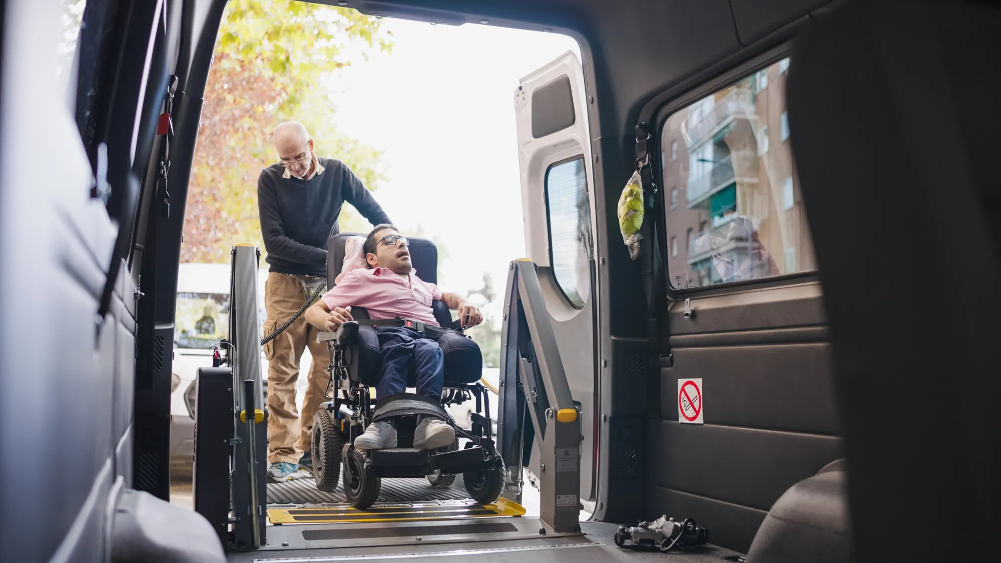 Senior man guiding a young man in a power wheelchair onto a van lift