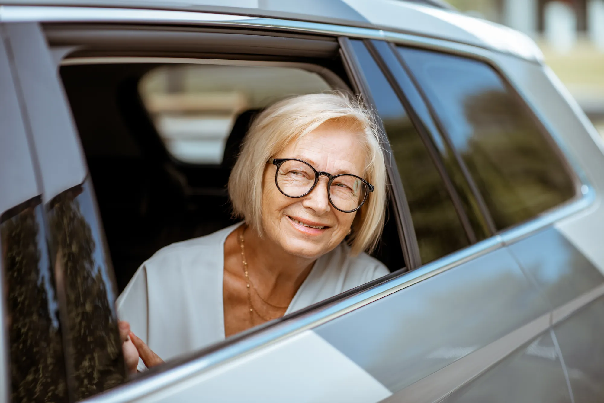 Senior woman in glasses smiling from the back seat window of a car